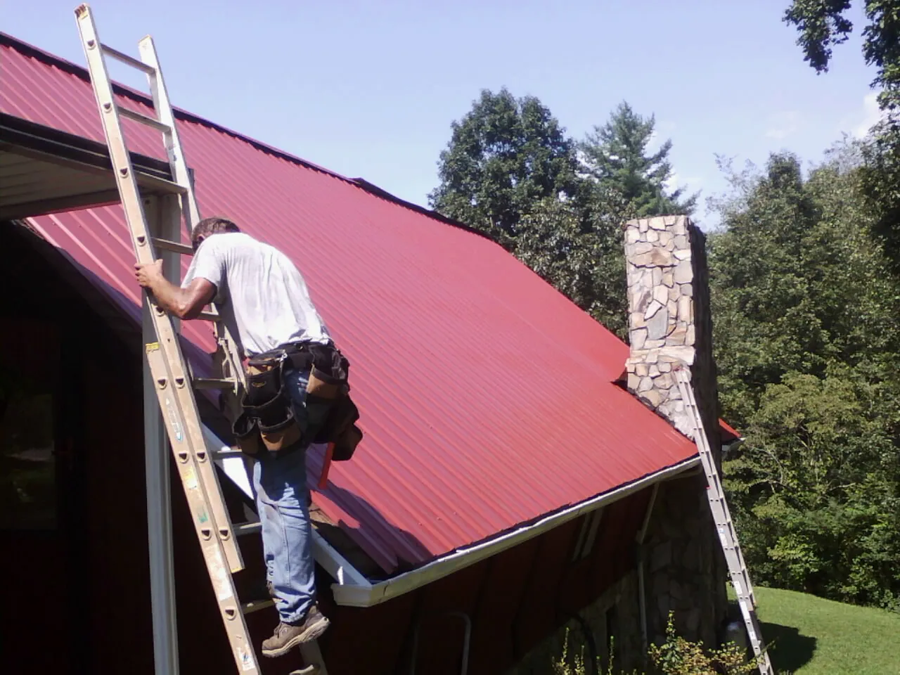 Roofer on a ladder beside a red metal roof and stone chimney — Soffit & Fascia Repair by Roofing Brothers in Woodmere